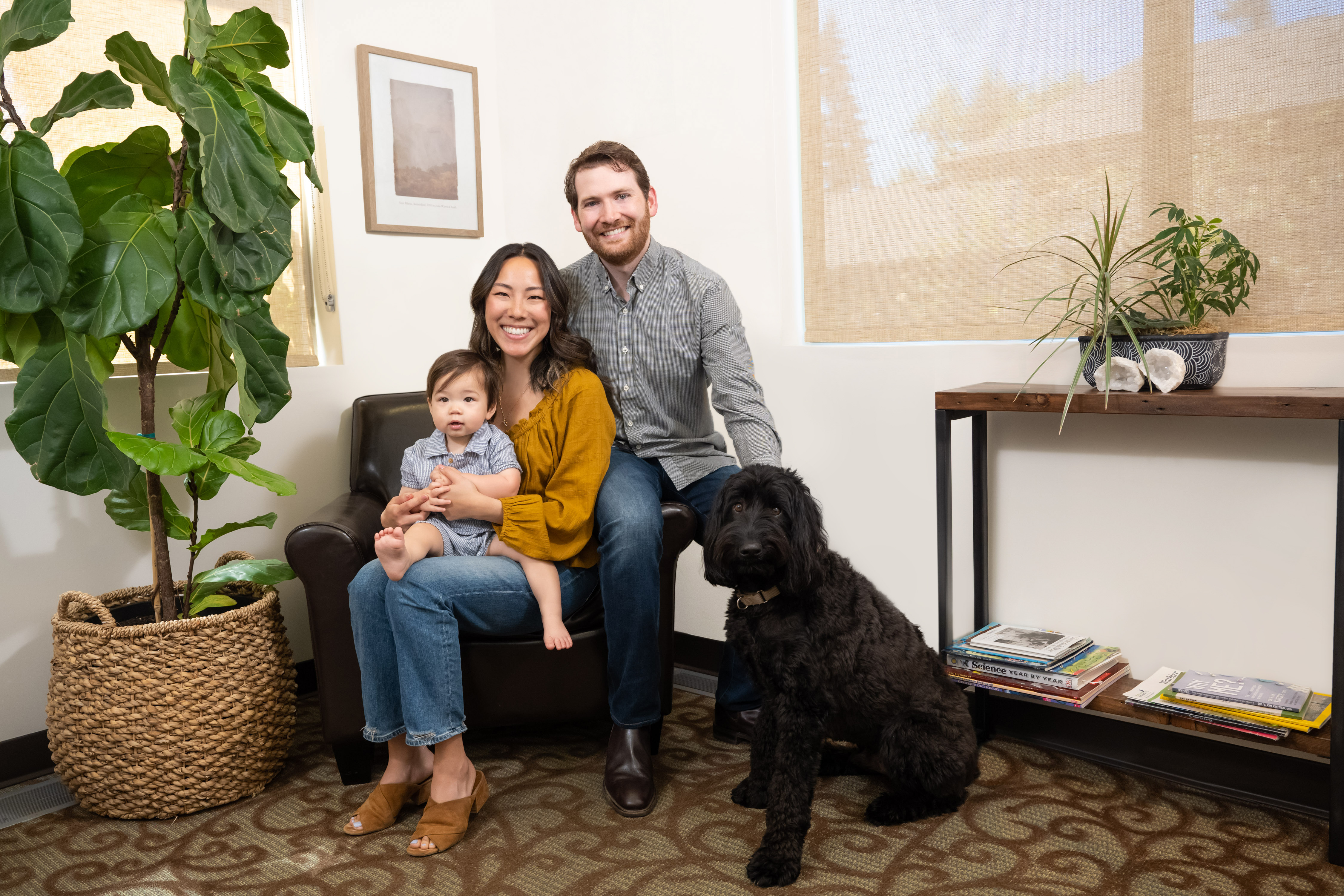A family of three posing in a room with a dog and potted plants, the man is seated on a couch while the woman stands beside him.