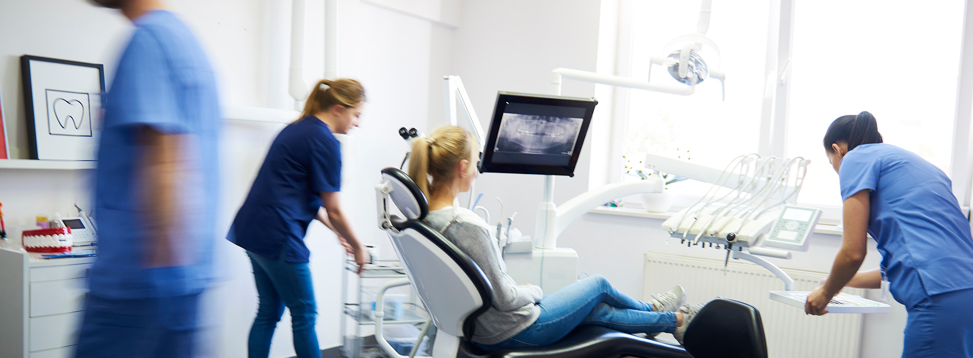 A group of healthcare professionals in a modern dental office, with one individual seated on an examination chair and another standing by a computer screen.