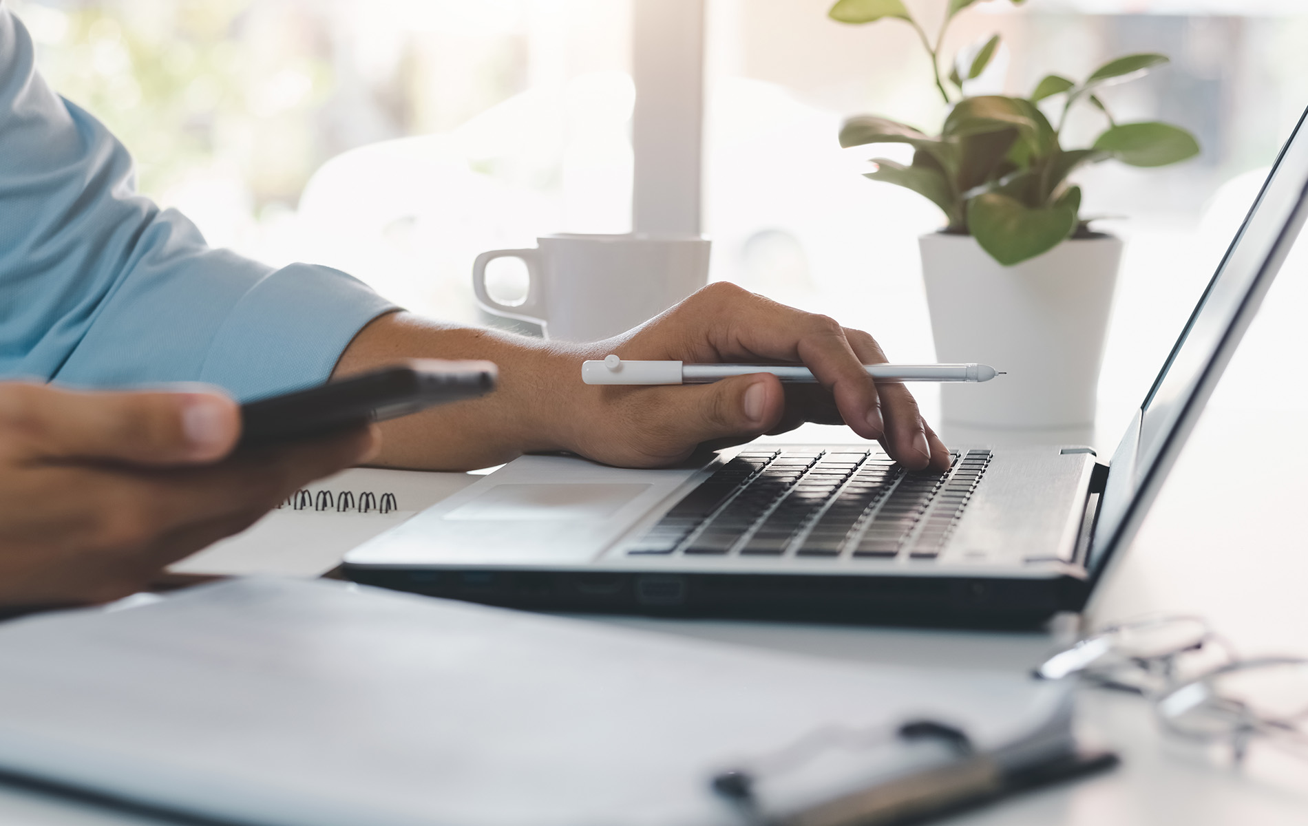 The image shows a person using a laptop at a desk with a handbag and a potted plant in the background.