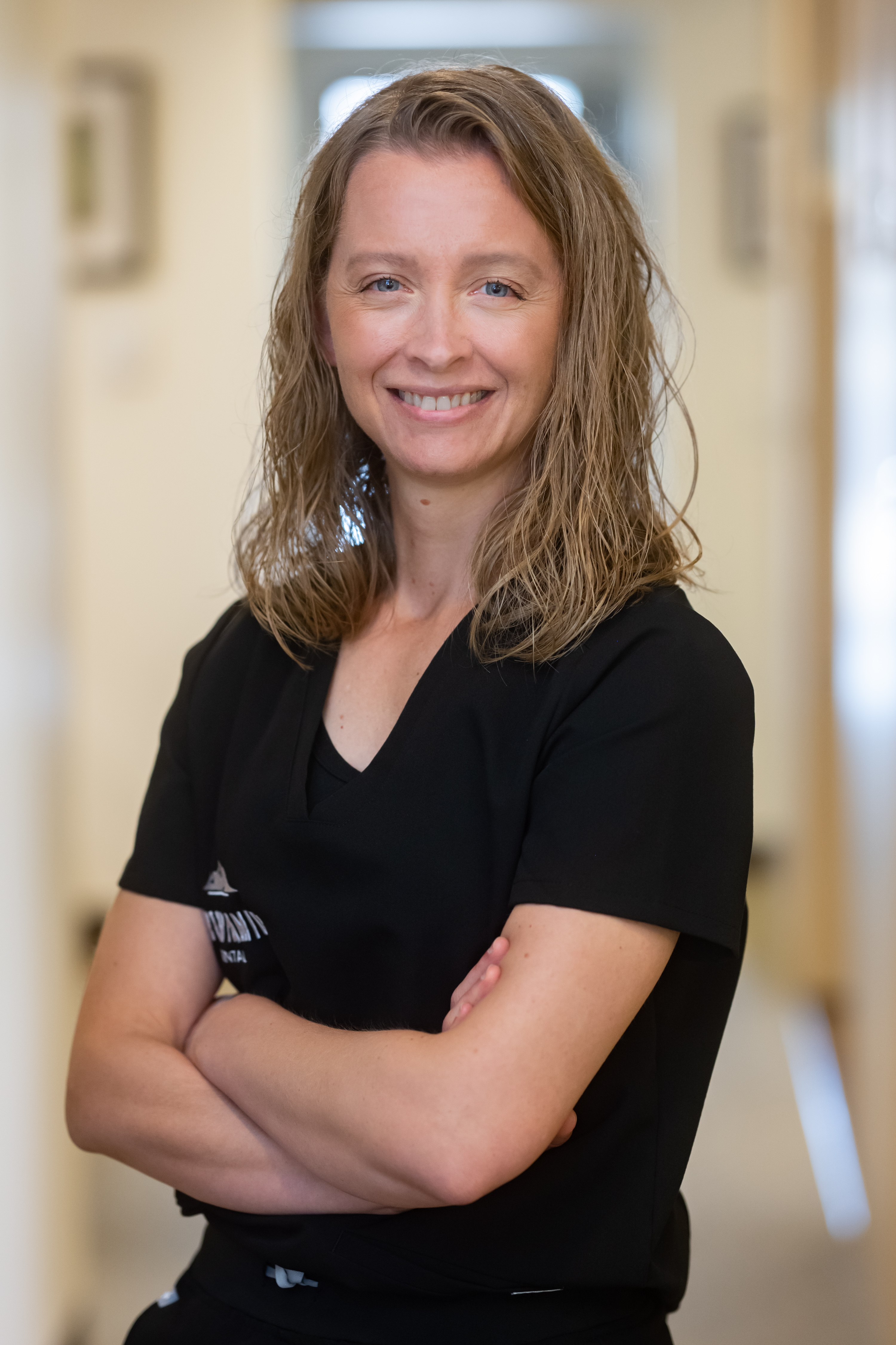 The image features a woman standing in an indoor setting, smiling at the camera. She is wearing a dark top and has her arms crossed. Her hair is styled short, and she appears to be posing for the photograph.