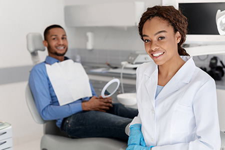 A person wearing a white lab coat stands in front of a dental chair, smiling at the camera, with another individual seated in the chair.