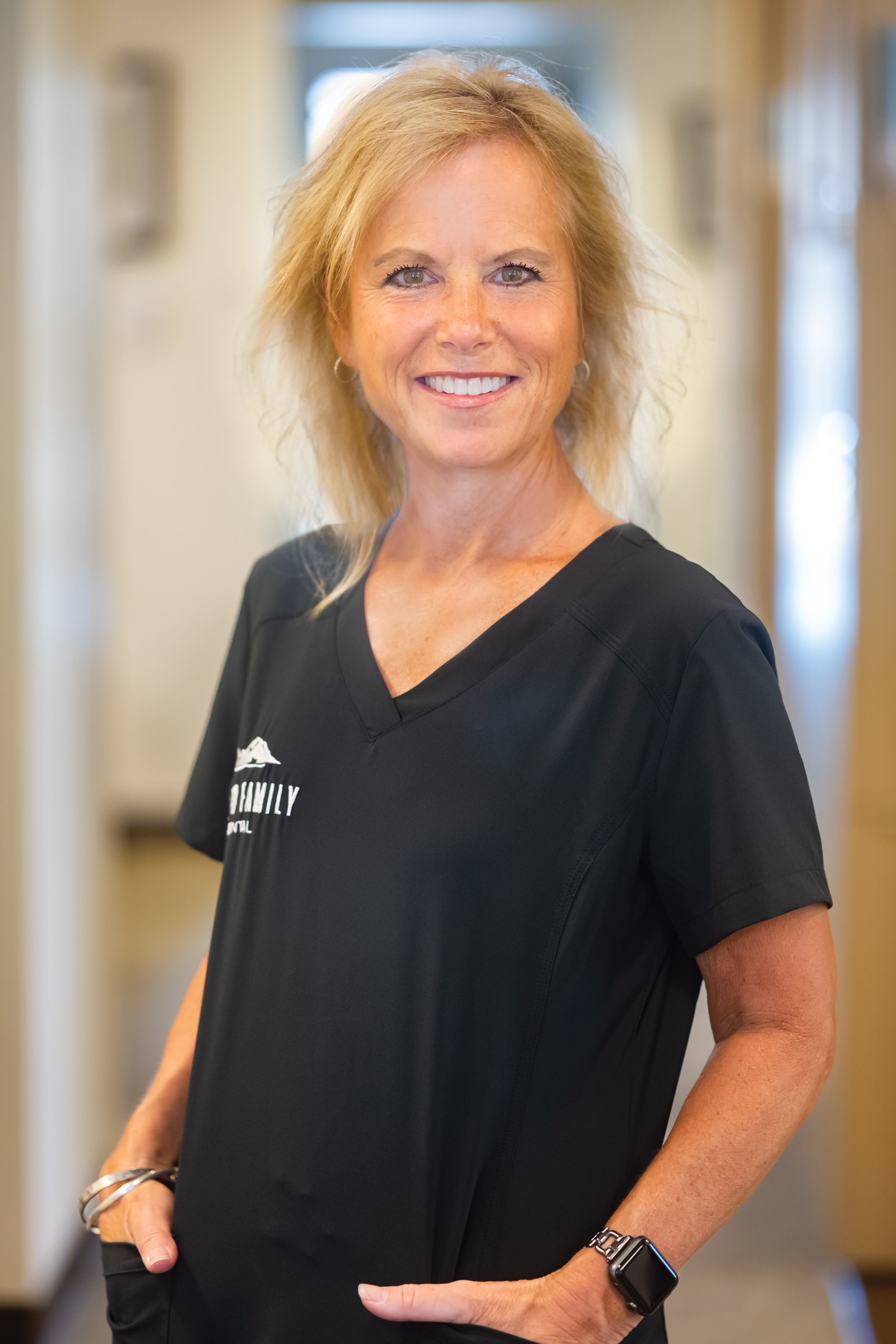 The image shows a woman standing in an office environment, wearing a black polo shirt with a logo on the left chest area and a name tag that is not fully visible. She has blonde hair styled in a short bob, and she's smiling at the camera. Her posture is relaxed, with her hands casually tucked into her pockets. The background suggests an indoor setting with a neutral color palette.