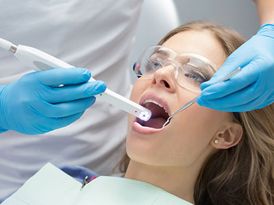 A dental professional using a device to clean or examine a patient s teeth, with the patient sitting in a chair and wearing blue gloves.